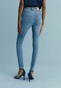 Jeans de mezclilla azul ajustados con cinco bolsillos, cintura alta y ligera elasticidad. Combinados con tacones abiertos oscuros. Textura suave, diseño clásico.