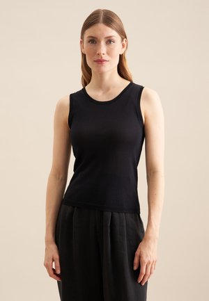 Vrouw die recht vooruit kijkt, draagt een mouwloos zwart topje en losse zwarte broek tegen een effen beige achtergrond.