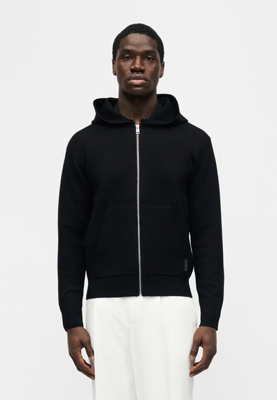 Νεαρός άνδρας που φοράει μαύρο φούτερ με φερμουάρ, κουκούλα και μπροστινές τσέπες, συνδυασμένο με λευκό παντελόνι, στέκεται μπροστά σε απλό ανοιχτό φόντο.