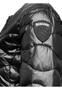 Zwarte pufferjack met een gequiltde textuur, glanzende stof en een prominente zwart schildvormige patch met het "Blauer"-logo.