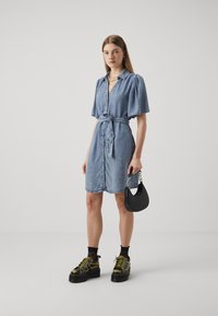 Vestido de camisa em denim com mangas curtas em balão, frente com botões e cintura com cinto. Combinado com sapatos de plataforma pretos e uma pequena bolsa preta.