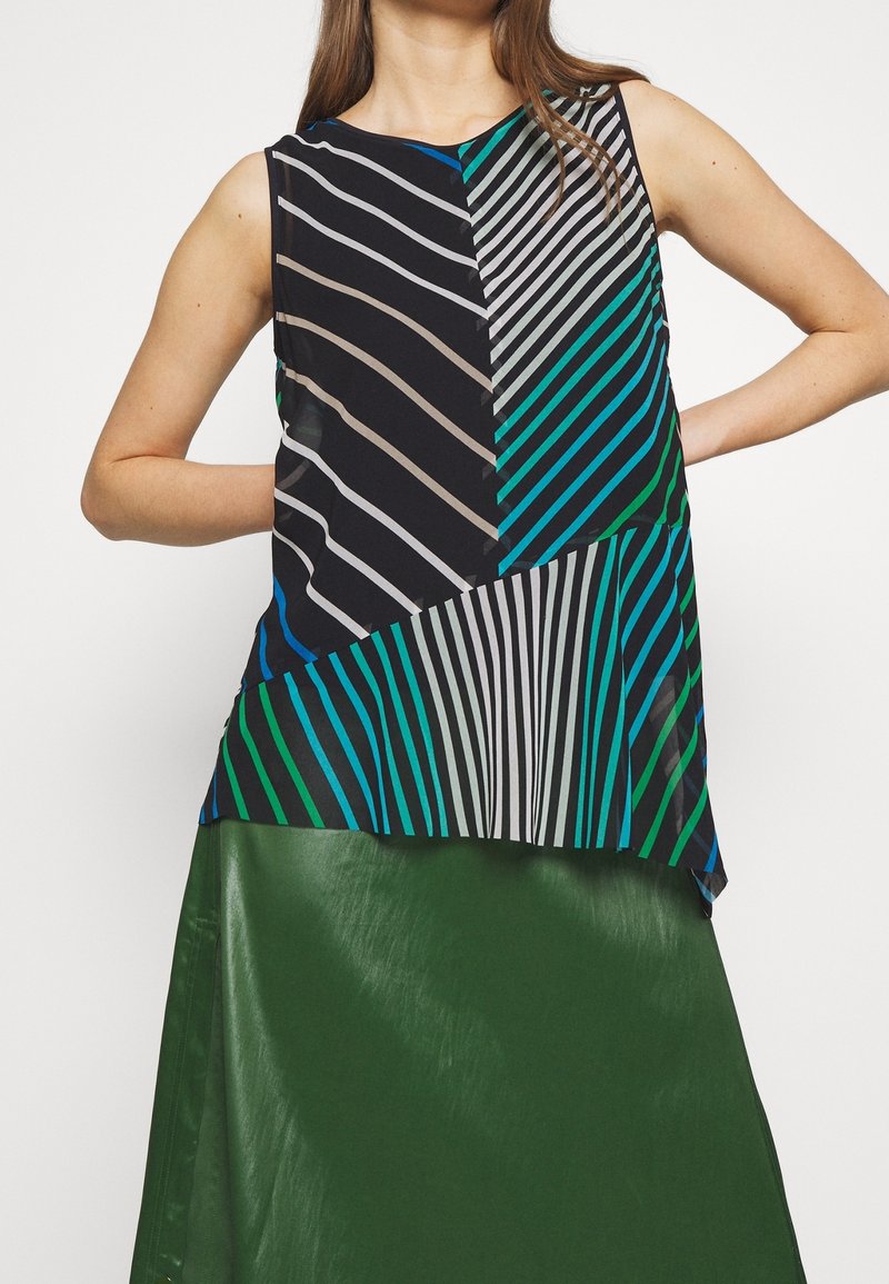 Haut sans manches à rayures multicolores diagonales, associé à une jupe vert foncé brillante, porté par une personne les mains sur les hanches.