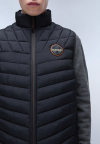 Schwarze gepolsterte Weste mit horizontalem Steppmuster, hohem Kragen, Frontreißverschluss und einem orangefarbenen, geprägten Logo-Patch auf der linken Brust.