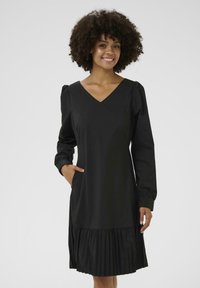 Robe noire avec un col en V, de longues manches bouffantes et un ourlet plissé. Fabriquée en tissu lisse avec des poches latérales, design minimaliste.