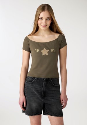 Giovane donna con lunghi capelli biondi indossa un top verde oliva scollato con stella beige e stampa "1991", abbinato a pantaloncini di jeans neri.