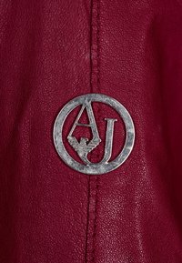 Tief burgunderfarbenes Leder mit einer glatten Textur, ausgestattet mit einem kreisförmigen silbernen Logo, in das die Buchstaben 'A' und 'J' in das Design integriert sind.