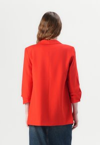 Czerwony żakiet z kołnierzykiem, długimi rękawami podwiniętymi na mankietach oraz prostym krojem. Gładki materiał i bezszwowa konstrukcja tyłu.