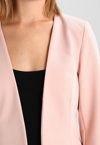 Blush pink blazer with textured fabric, open front, and no fastenings. Worn over a black top, showcasing a simple and modern design.
