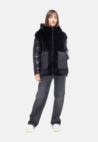 Giacca in pelliccia sintetica nera con tessuto lucido imbottito nero, chiusura con zip, tasche frontali e cappuccio. Indossata sopra jeans grigio scuro e sneaker.
