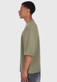 T-shirt oversize verde oliva realizzato in morbido tessuto, caratterizzato da maniche corte e scollatura rotonda. Include dettagli di cucitura sottili.
