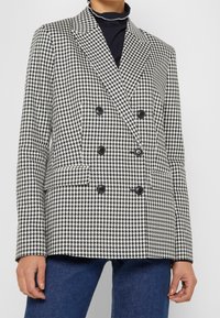 Vrouw in een zwart-wit houndstooth dubbelzijdig colbert met zwarte knopen, gecombineerd met donkerblauwe jeans en een zwarte coltrui.
