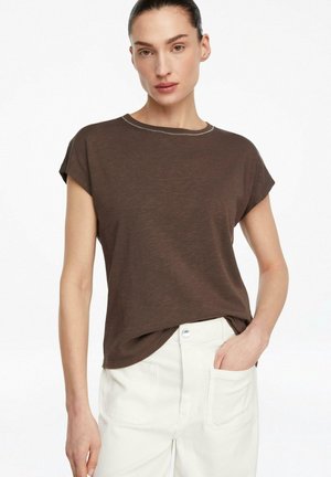 Donna che indossa una maglietta marrone a maniche corte e pantaloni bianchi, con una mano in tasca, in piedi davanti a uno sfondo bianco semplice.