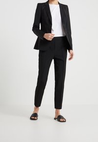 Svart skräddarsydd kavaj med en enkel knäppning, tillsammans med slim-fit svarta byxor och svarta platta sandaler. Den vita t-shirten finns under.