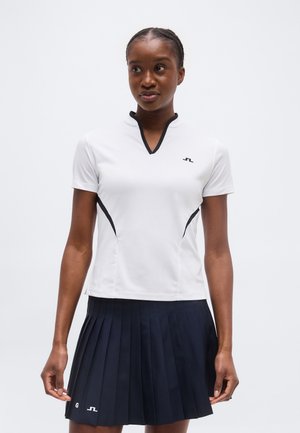 Γυναίκα που φοράει λευκό τοπ με κοντά μανίκια και μαύρο περίγραμμα, και φούστα σε μπλε ναυτικό χρώμα με πτυχώσεις, στέκεται μπροστά σε λευκό φόντο.