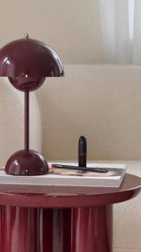 Bordeaux bordlampe med en rund skærm, et blankt bordeaux bord og et magasin. Sort læbestift og liner placeret på magasinet.
