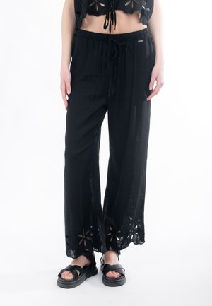 Schwarze, leichte, weit geschnittene Hose mit Kordelzug am Bund. Verziert mit floralen Cut-out-Details am Saum und aus strukturiertem Stoff. Schwarze Sandalen.