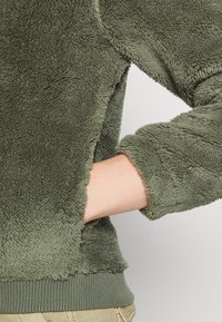 Polarowy pulower w stonowanej zieleni, z miękkim, teksturowanym wykończeniem, ściągaczami przy rękawach oraz luźnym krojem, z boczną kieszenią na rękę.