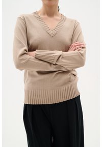 Beige stickad tröja med V-ringning som har ribbade detaljer runt halsringningen och ärmarna. Nederkanten är också ribbad, vilket ger en texturerad finish.