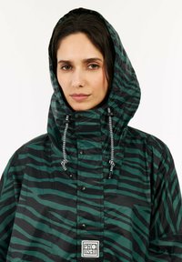 Zielono-czarny, paskowany płaszcz przeciwdeszczowy z kapturem, posiadający sznurki ściągające, zatrzaski oraz małe logo na dole. Materiał wodoodporny.