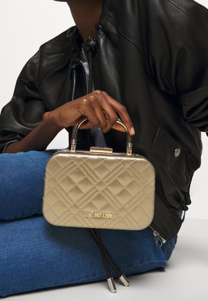 Kulttuurimaton kultainen käsilaukku suorakulmaisen muotoisena, metallikahvoilla ja brändilogoilla. Ominaisuuksia kiiltävä pinta ja erottuvat tummat hihnat.