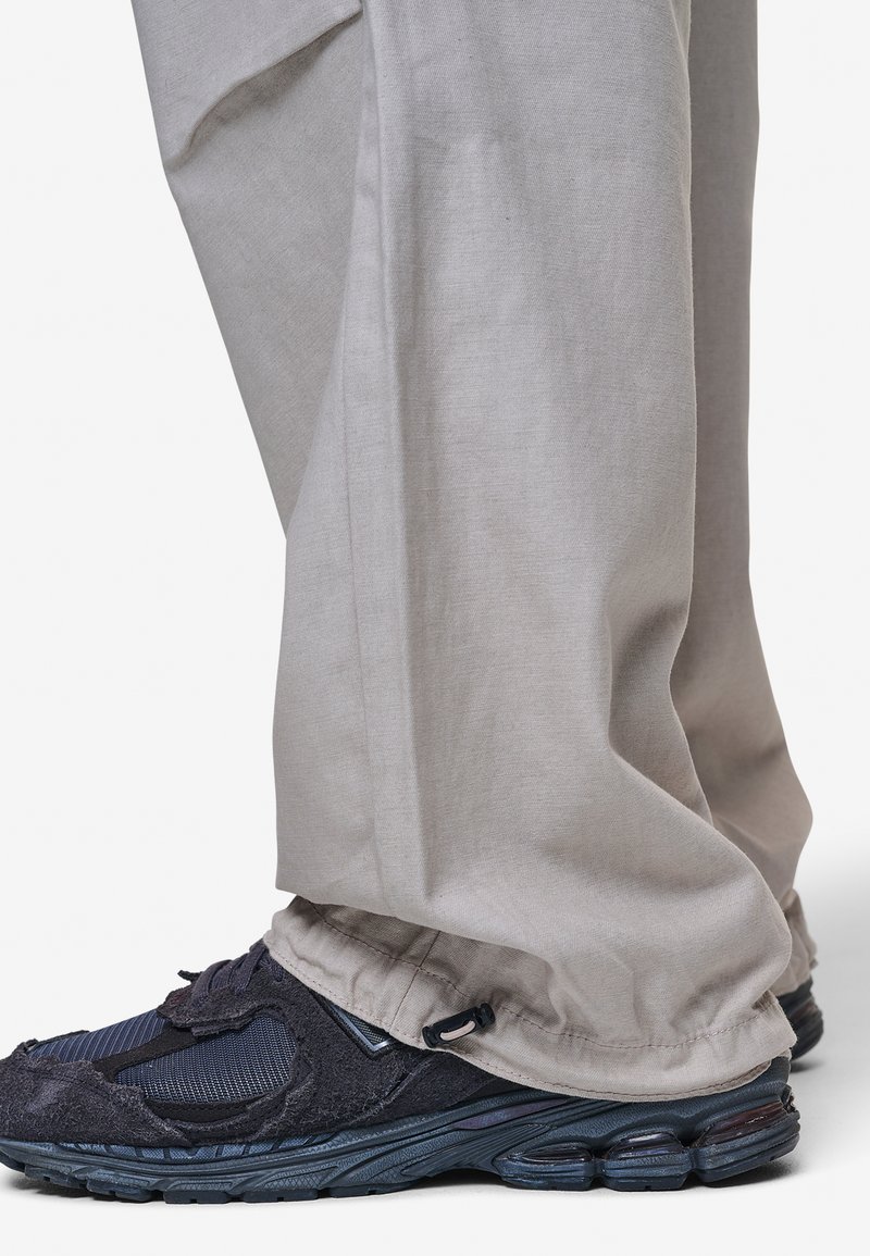 Luźne beżowe spodnie zebrane w kostce, noszone z czarnymi i granatowymi butami sportowymi o teksturowanej tkaninie i wyściełanych podeszwach.