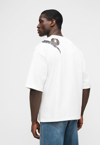 Άνδρας που φοράει λευκό υπερμεγέθη μπλουζάκι με μπλε και καφέ σχέδιο φιδιού στον ώμο, συνδυασμένο με μπλε τζιν, κοιτάζοντας πλάγια.