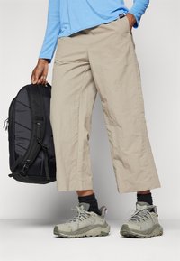 Beige cropped pants with a textured finish, paired with gray hiking shoes and a black backpack. Blue long-sleeve shirt visible.