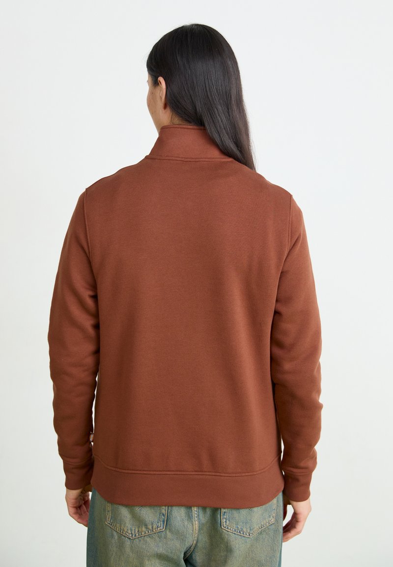 Felpa marrone con colletto alto, texture morbida, polsini e orlo a coste. Indossata sopra jeans azzurri chiari, con dettagli di cucitura visibili sul retro.