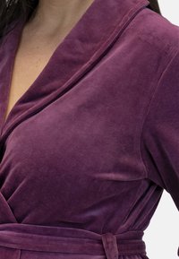 Accappatoio viola in velluto con collare a scialle, dotato di una vita legata e di una consistenza morbida. Il tessuto ha una leggera lucentezza e un aspetto liscio.