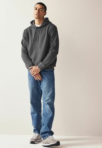 Grå tekstureret hoodie med snoretræk, parret med blå denim jeans og grå sneakers med en hvid accent. Står foran en neutral baggrund.