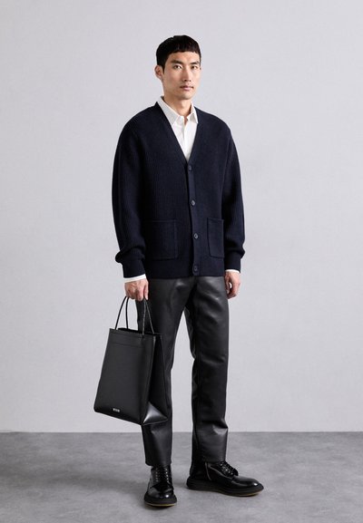 Mees, kes kannab tumesinist kardigani, valget särk, musti nahkpükse ja saapaid, hoiab käes musta kottu, taustal on hall ühtlane taust.