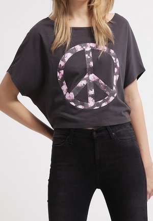 Femme portant un T-shirt court noir avec un signe de paix floral rose, associé à un jean noir taille haute, debout avec une main sur la hanche.