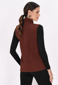 Vest din fleece maro cu guler înalt. Purtați peste o cămașă neagră cu mânecă lungă. Textură netedă, design ajustat și o croială simplă, curată.
