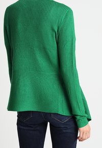 Grön ribbad stickad cardigan med långa ärmar, med en utflärdad nederkant och textilstruktur; bärs över mörkblå jeans.