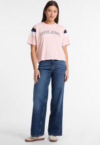 Ružový cropped tričko s logom 'Guess Jeans', krátke rukávy a námornícke akcenty. Nosené s širokými modrými džínami a pohodlnými teniskami.