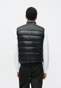 Čierna prešívaná vesta s lesklým povrchom, vybavená horizontálnymi prešívanými časťami, vysokým golierom a bez rukávov, nosená cez svetlý sveter.