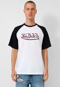 Bijela majica s crnim raglan rukavima, s „Von Dutch“ logotipom u tamnocrvenoj boji preko prsa. Izrađena od mekog pamučnog materijala.