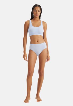 Donna con i capelli intrecciati che indossa un reggiseno sportivo senza cuciture azzurro chiaro e biancheria intima a vita alta, in piedi a piedi nudi contro uno sfondo bianco.