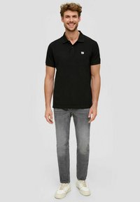Schwarzes Poloshirt mit einem kragenförmigen Design, kurzen Ärmeln und einem kleinen gestickten Logo auf der Brust, kombiniert mit hellgrauen Jeans und Sneakers.