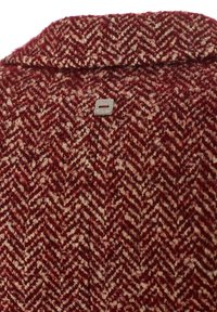 Tessuto rosso a motivo a spina di pesce con una texture morbida. Presenta un piccolo tag in metallo con una fessura, che arricchisce il dettaglio del design.