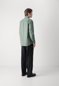 Camisa de botón verde claro con mangas largas, corte holgado, dobladillo redondeado, combinada con pantalones anchos oscuros y zapatos negros.