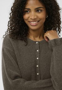 Nasmejana ženska s kodrastimi lasmi nosi mehak rjav cardigan s perlastimi gumbi, nežno drži ovratnik z eno roko.