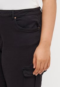 Svarta cargobyxor med sidofickor, slim fit och slät textur. Synlig sömnad och metalldetaljer nära fickan.