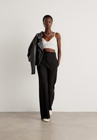 Schwarze hochgeschnittene Hosen, weißes, kurz geschnittenes Oberteil mit strukturierten Details und eine schwarze Lederjacke. Das Model posiert in einem minimalistischen Setting.