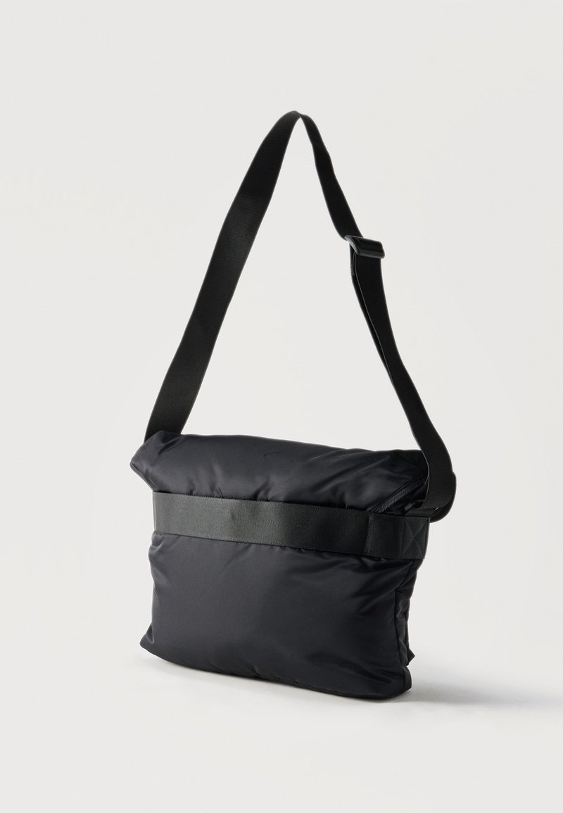 Sac à bandoulière noir matelassé avec sangle réglable et large bande horizontale sur le devant, présenté sur un fond clair uni.