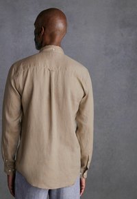 Heren overhemd met lange mouwen, van beige linnen materiaal, met kraag en een knoopsluiting aan de achterkant, gebogen zoom en zichtbare naaideetails. Textuurafwerking.