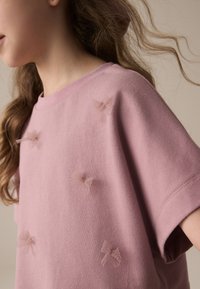 Lichtroze katoenen T-shirt met een ronde hals, korte mouwen en geborduurde tule strikjes op verschillende plekken aan de voorkant. Zachte textuur.