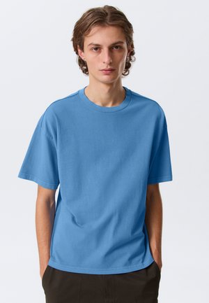 Jeune homme aux cheveux bruns mi-longs portant un t-shirt bleu uni et un pantalon foncé, debout devant un fond gris clair.