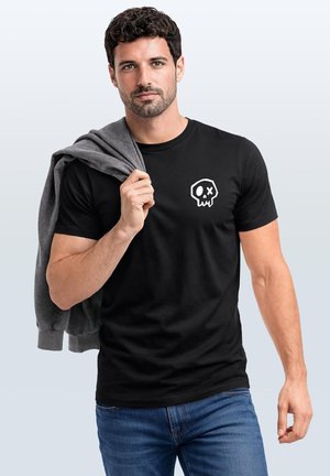 Junger Mann mit kurzen lockigen Haaren trägt ein schwarzes T-Shirt mit weißem Totenkopf-Logo und hält eine graue Jacke über der Schulter.