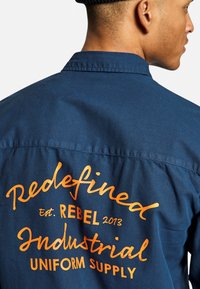 Camicia blu scuro con bottoni, con testo arancione stampato sul retro: "Ridefinito Est. REBEL 2013 FORNITURA UNIFORME INDUSTRIALE."